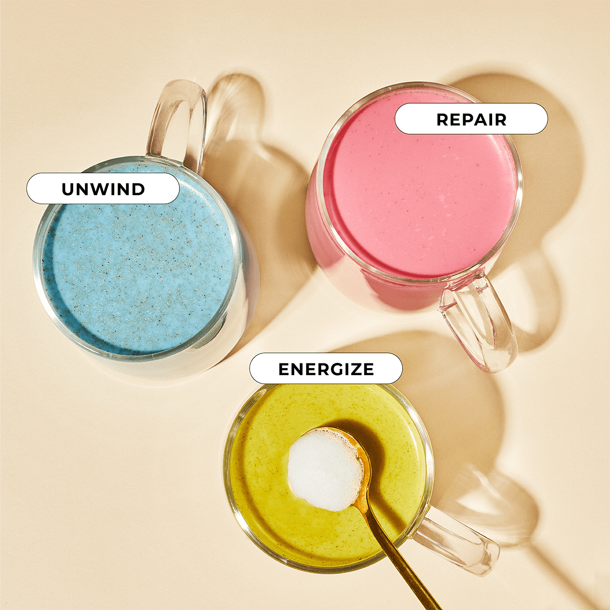 On a beige surface, three Blume glass mugs from The All Day Bundle serve distinct functions: UNWIND holds Blue Lavender liquid, REPAIR contains pink liquid, and ENERGIZE features yellow liquid with a spoon of white powder in it.